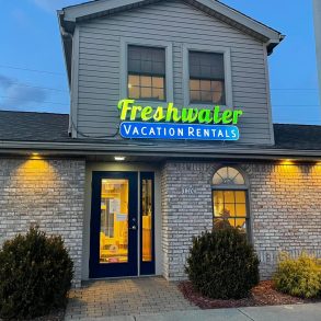 A sign reading Freshwater Vacation Rentals lit up hanging over an office with the lights on inside on a cloudy evening.