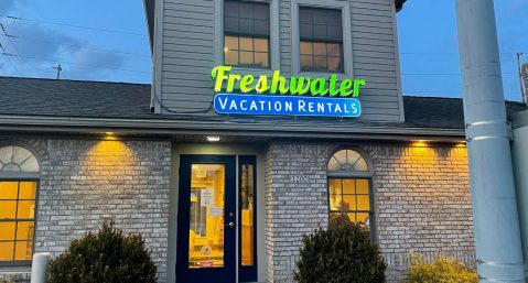 A sign reading Freshwater Vacation Rentals lit up hanging over an office with the lights on inside on a cloudy evening.