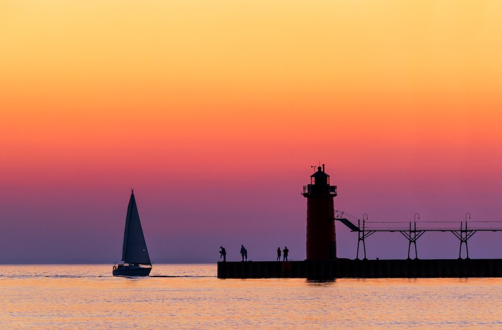 A vividly colorful twilight sky silhouettes a sailboat, people, and the lighthouse at South Haven, Michigan on Lake Michigan.