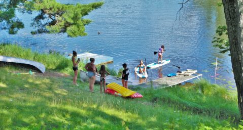 Parents watching children jump off a dock and swim in a lake.
